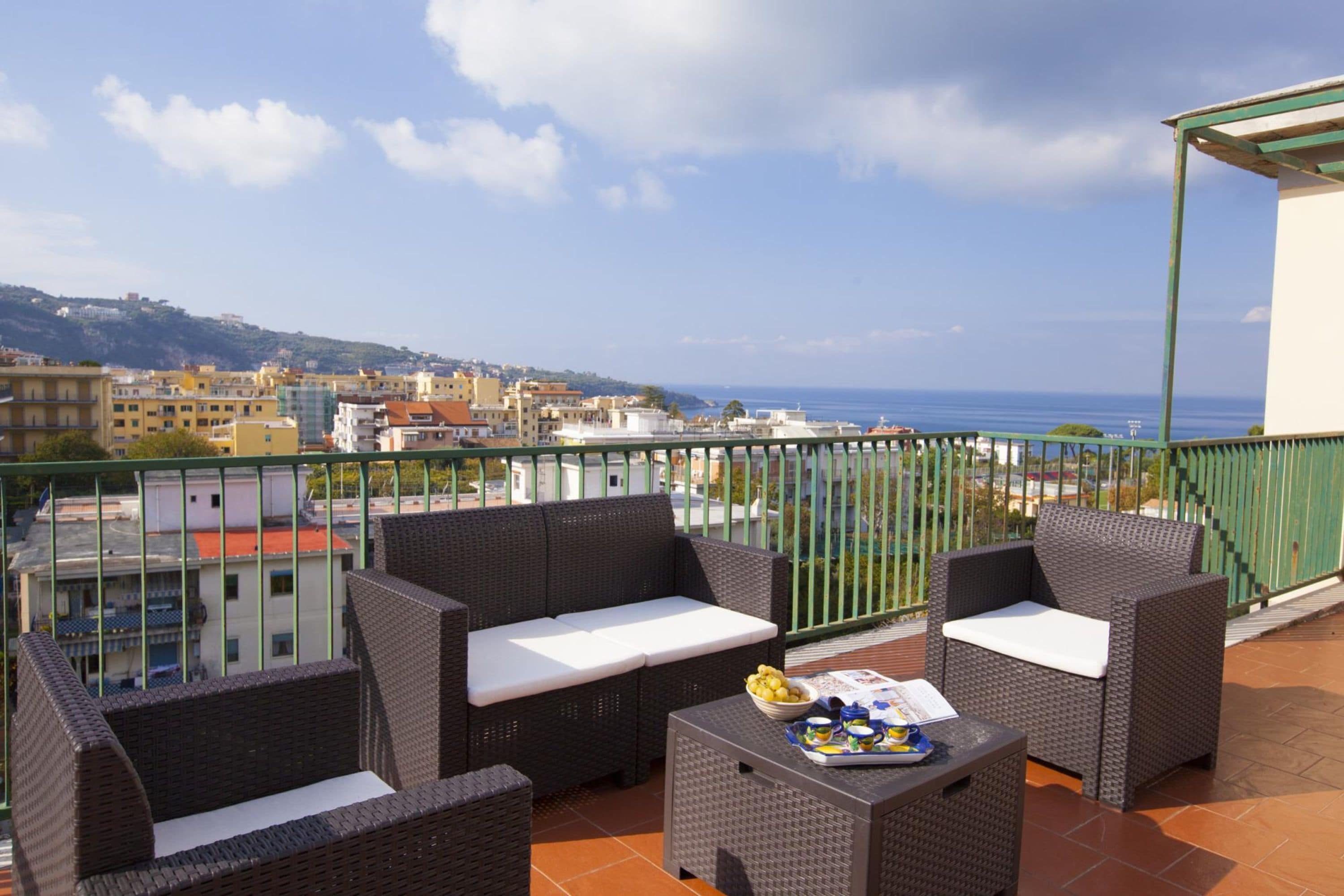 Ein Balkon mit Korbmöbeln, einem Tisch mit Obstschale und Blick auf das Meer und Gebäude.