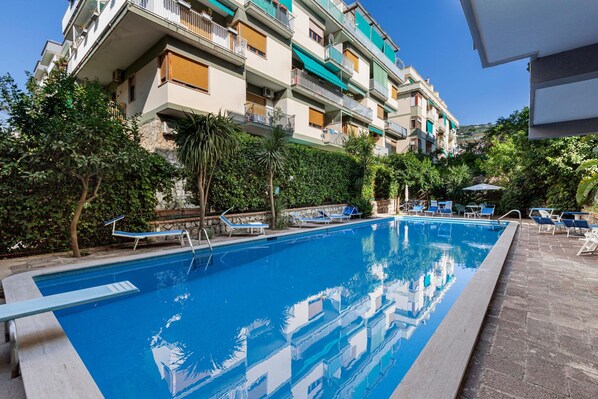 Une piscine entourée de chaises longues, elle-même bordée d’un immeuble à plusieurs étages doté de balcons et de verdure.