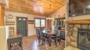 A dining area with a wooden table and chairs, a stone fireplace, and a kitchen area with cabinets and a window. - 3 Mi to Trails: Woodsy Retreat in Overgaard! (Overgaard)
