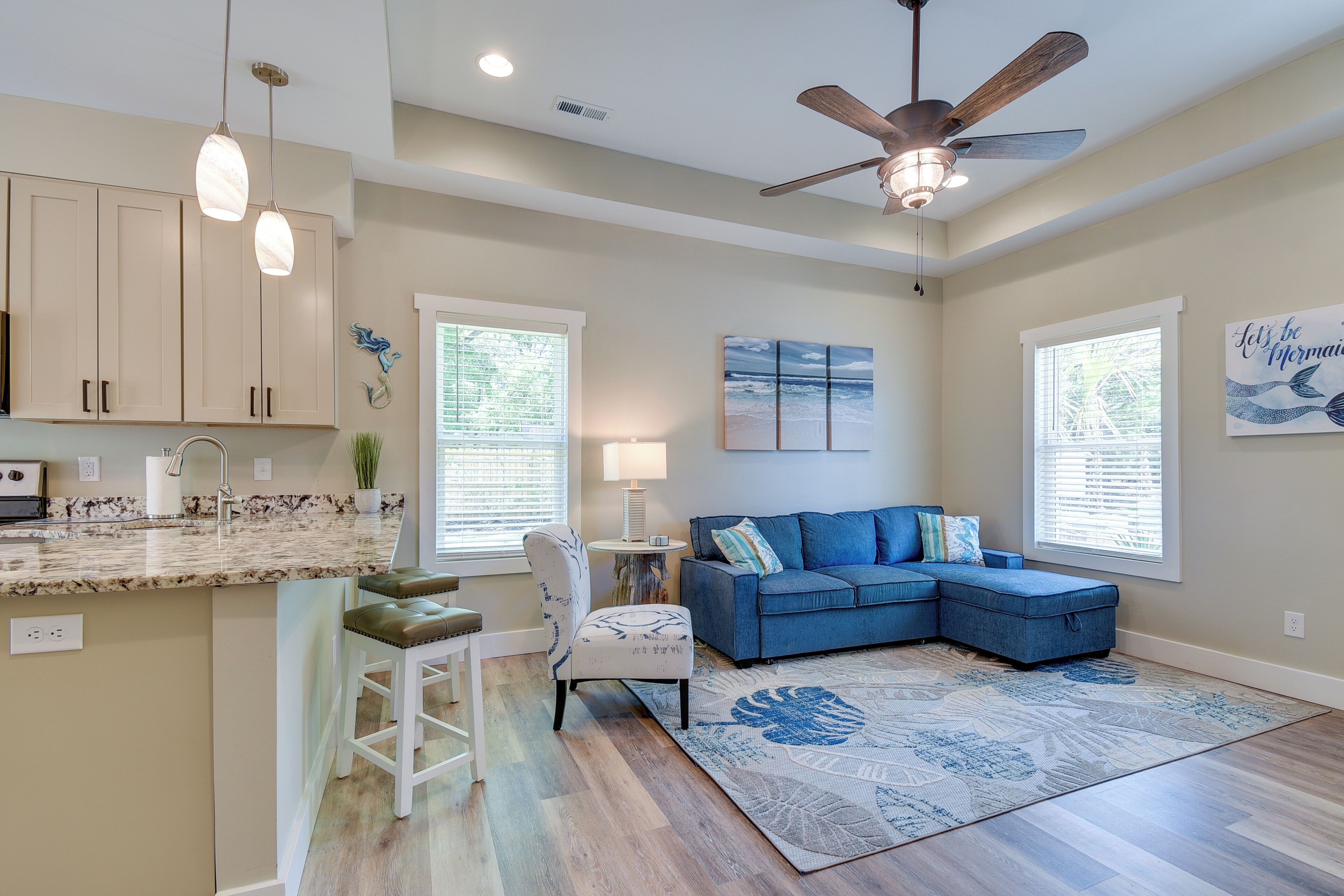 A spacious living area with a blue sectional sofa, a ceiling fan, and a kitchen with a marble countertop.