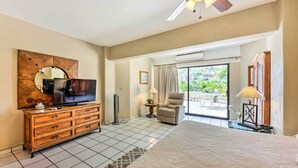 Habitación de hotel con una cama grande, un televisor en una cómoda de madera, una silla, una mesita con una lámpara y vistas al exterior por una puerta corredera.