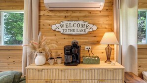 Ein Holzblockhaus-Innenraum mit einem 'WELCOME TO THE LAKE' Schild, einer Keurig-Kaffeemaschine, einer Lampe und einem Korbwäschekorb auf einer Holzkommode.