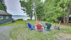 Eine grüne, offene Landschaft mit Seeblick, umgeben von Bäumen und mehreren Adirondack-Stühlen.