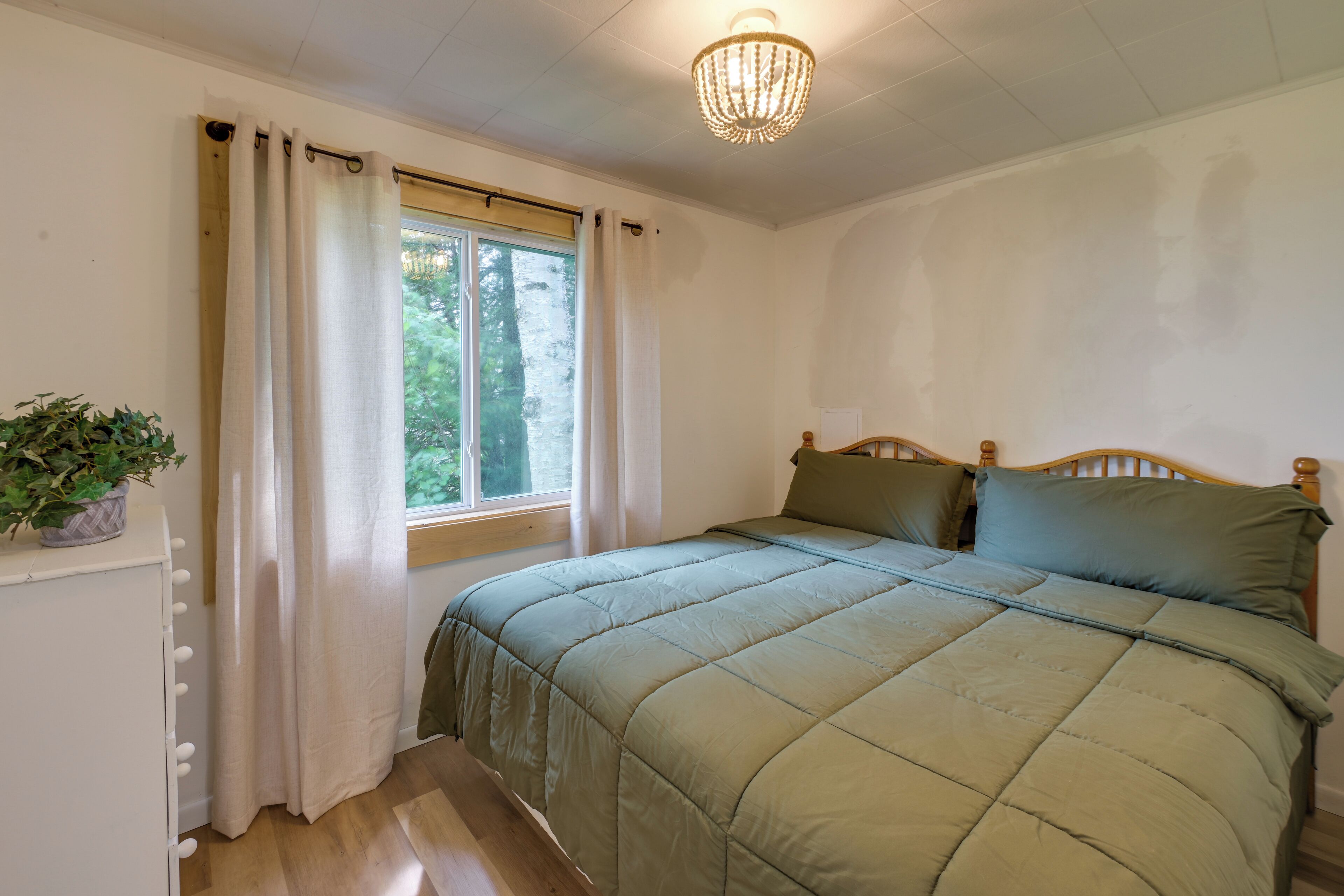Um quarto com cama, janela com cortinas, um candelabro e um vaso com planta.
