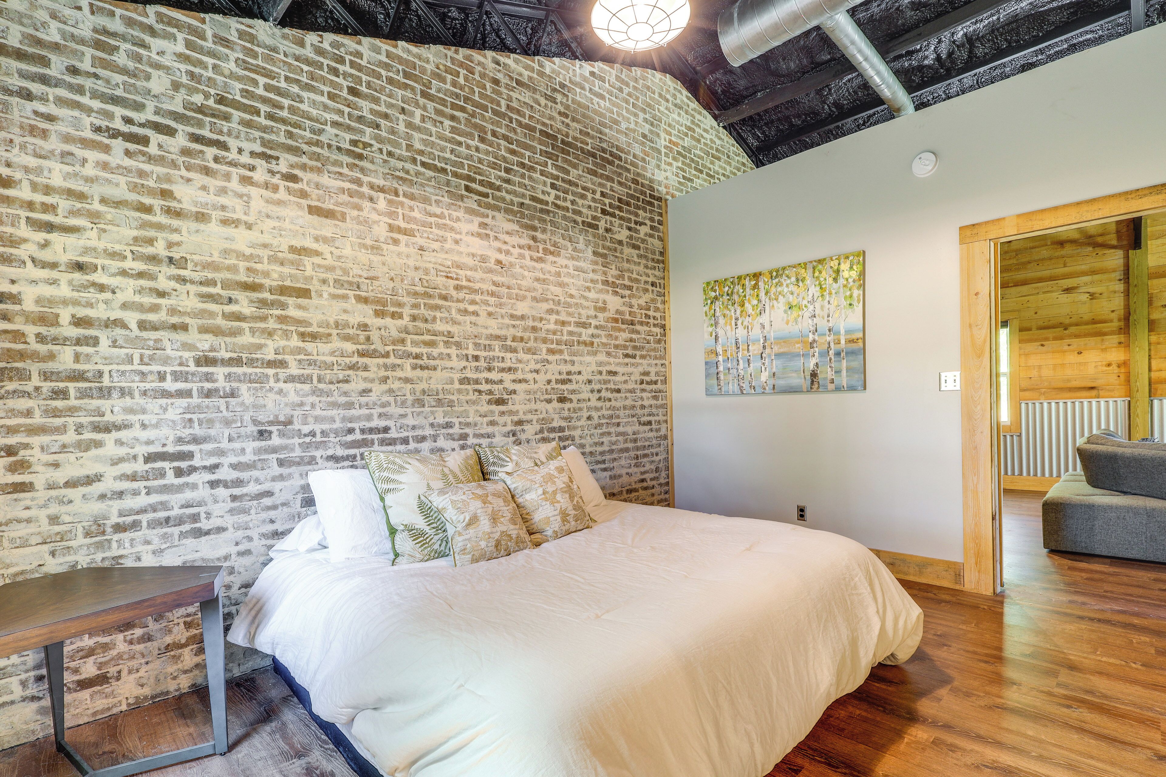 A bedroom with a brick wall, a bed with white bedding, a wooden bench, and a painting on the wall.