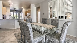 A modern kitchen with a central island, a dining area with patterned chairs, and a large window with blinds.