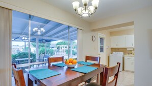 Un coin repas avec une table en bois, des chaises, un saladier rempli de fruits et une vue sur l’extérieur grâce à de grandes fenêtres.