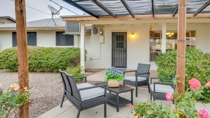 Un espace patio aménagé avec des meubles en osier et une table, entouré de verdure et avec une maison en arrière-plan.