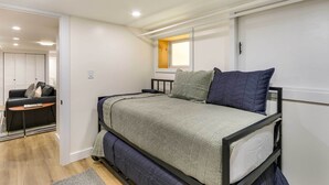 Quarto com cama de solteiro e beliche, uma mesinha, um sofá e janela com vista para o exterior.