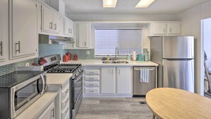 Une cuisine moderne avec des armoires blanches, des appareils en acier inoxydable, un four à micro-ondes, une cuisinière et un lave-vaisselle. Il y a une fenêtre avec des stores et une table en bois.