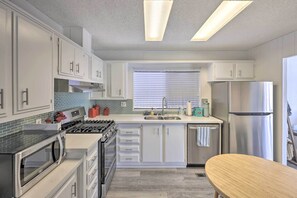Una cocina moderna con armarios blancos, electrodomésticos de acero inoxidable, microondas, estufa y lavavajillas. Hay una ventana con persianas y una mesa de madera.
