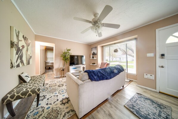A living room with a sofa, bench, ceiling fan, TV, and a large window. - Pet-friendly Mayfield Heights Vacation Rental! (Mayfield Heights)