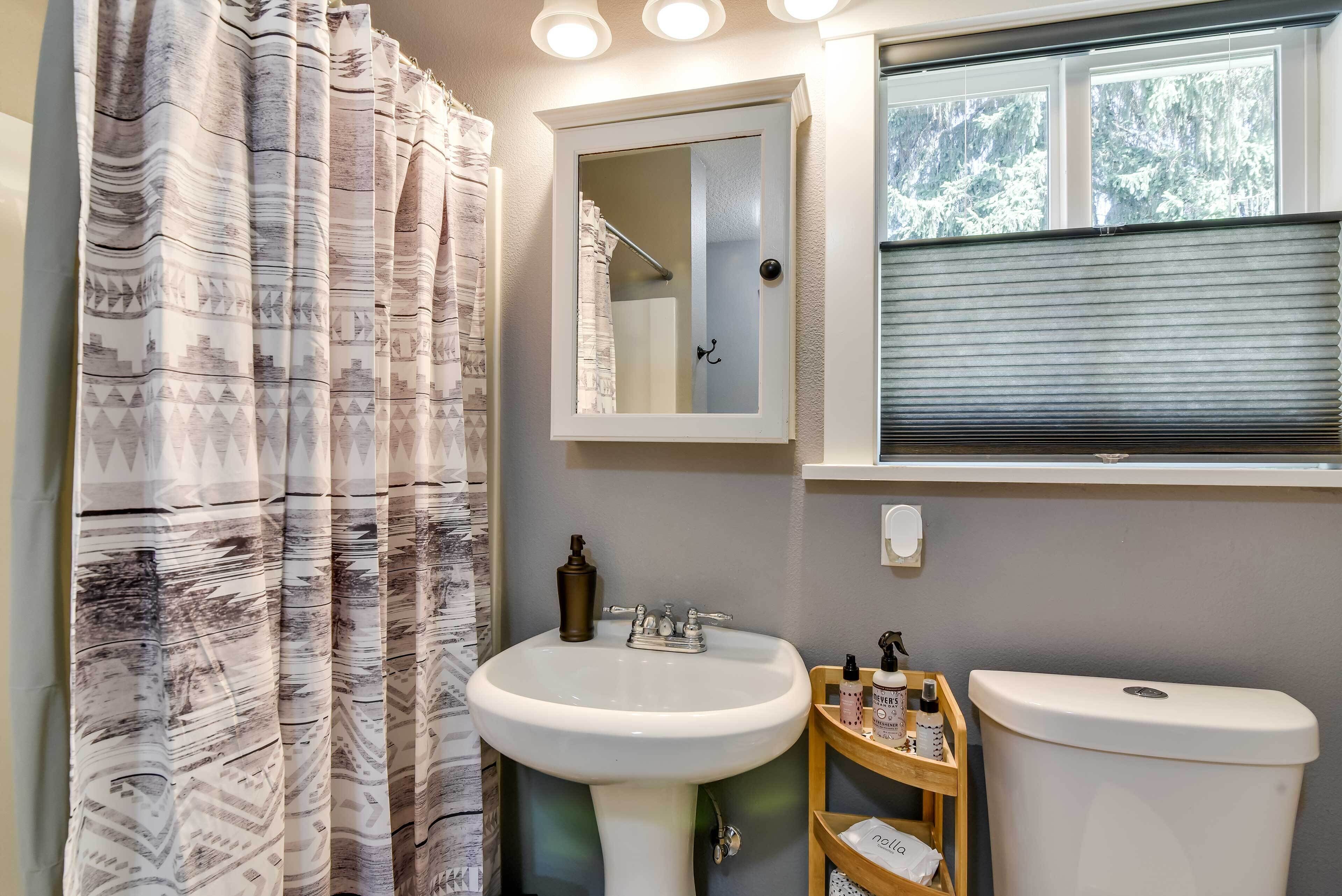 A bathroom with a white sink, a mirror above it, and a toilet. There is a window with blinds, and a shower curtain with a geometric pattern.