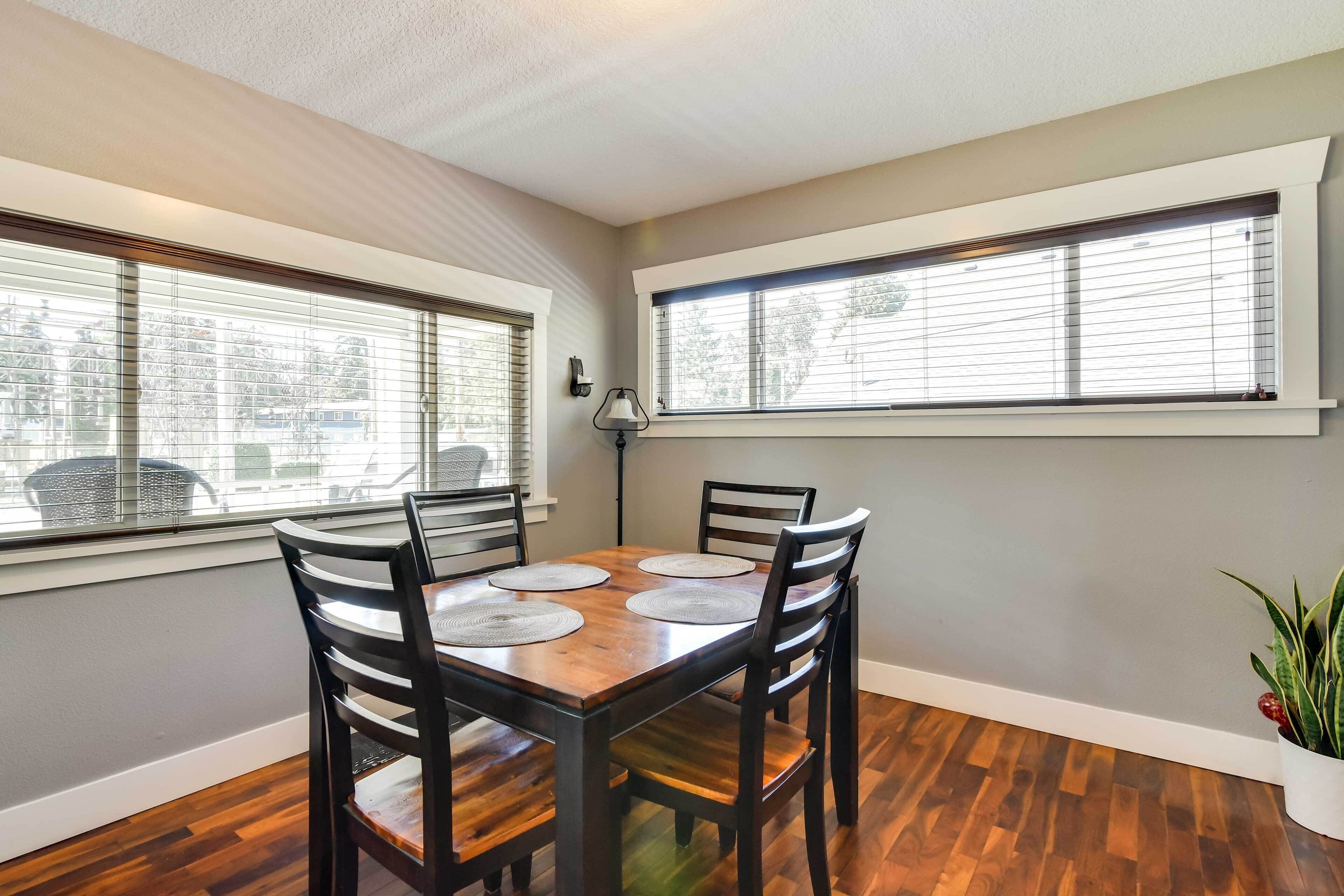 A dining area with a wooden table and four chairs, large windows with blinds, and a potted plant on the floor.
