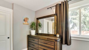 Une pièce comprenant une commode en bois, un grand miroir, une plante en pot et une fenêtre avec des rideaux.