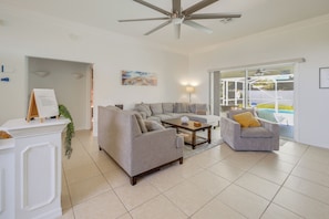 A modern living room with a grey sofa, a coffee table, and a grey armchair. There is a ceiling fan, a painting on the wall, and a view of the outside through a glass door.