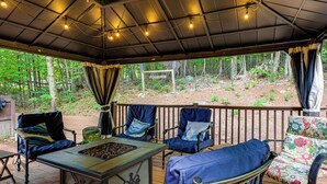 An outdoor patio with a wooden deck, metal-framed chairs, a metal table with a tray of food, and a view of a forested area. - Rustic Adirondacks Home w/ Hot Tub & Lake Access! (Chestertown)