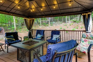 An outdoor patio with a wooden deck, metal-framed chairs, a metal table with a tray of food, and a view of a forested area. - Rustic Adirondacks Home w/ Hot Tub & Lake Access! (Chestertown)