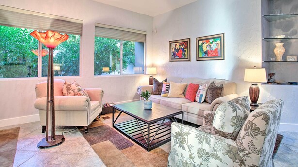 A living room with a sofa, armchairs, a coffee table, and a floor lamp. There are two windows with blinds, a painting on the wall, and a shelf with decorative items. - Spacious Palm Desert Retreat w/ Outdoor Oasis! (Palm Desert)