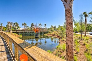 Une promenade en bois bordant un plan d’eau paisible, avec des palmiers et un pont en arrière-plan.