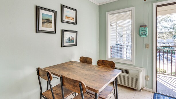 A dining area with a wooden table and four chairs, framed pictures on the wall, a window with blinds, and a small decorative item. - Pool & Beach Access: Hilton Head Coastal Condo (Hilton Head Island)