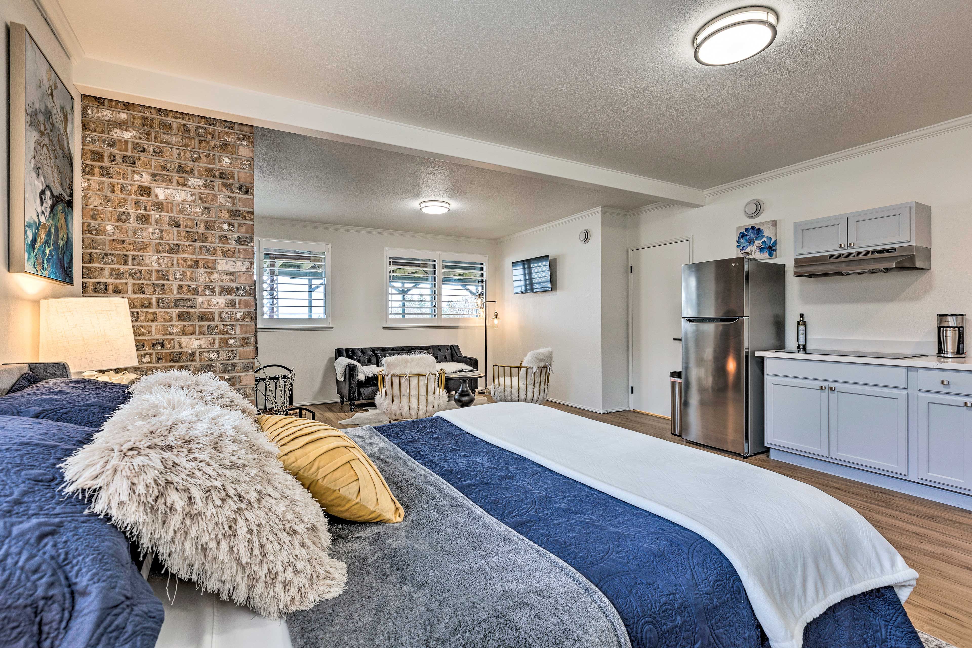 A modern bedroom with a large bed, a brick accent wall, a kitchenette, and a view of the living area.