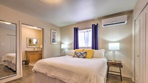Une chambre à coucher avec un grand lit, des tables de chevet, un appareil de climatisation et une salle de bain visible à travers une porte ouverte.