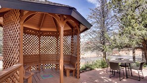 Ein hölzerner Gazebo mit Gitterwänden und Bänken, umgeben von einer Terrasse mit Tisch und Stühlen.