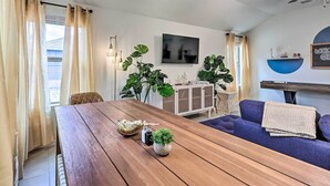 A modern living room with a wooden dining table, blue sofa, and large green plants. - Modern Granite Shoals Retreat w/ Fire Pit! (Marble Falls)