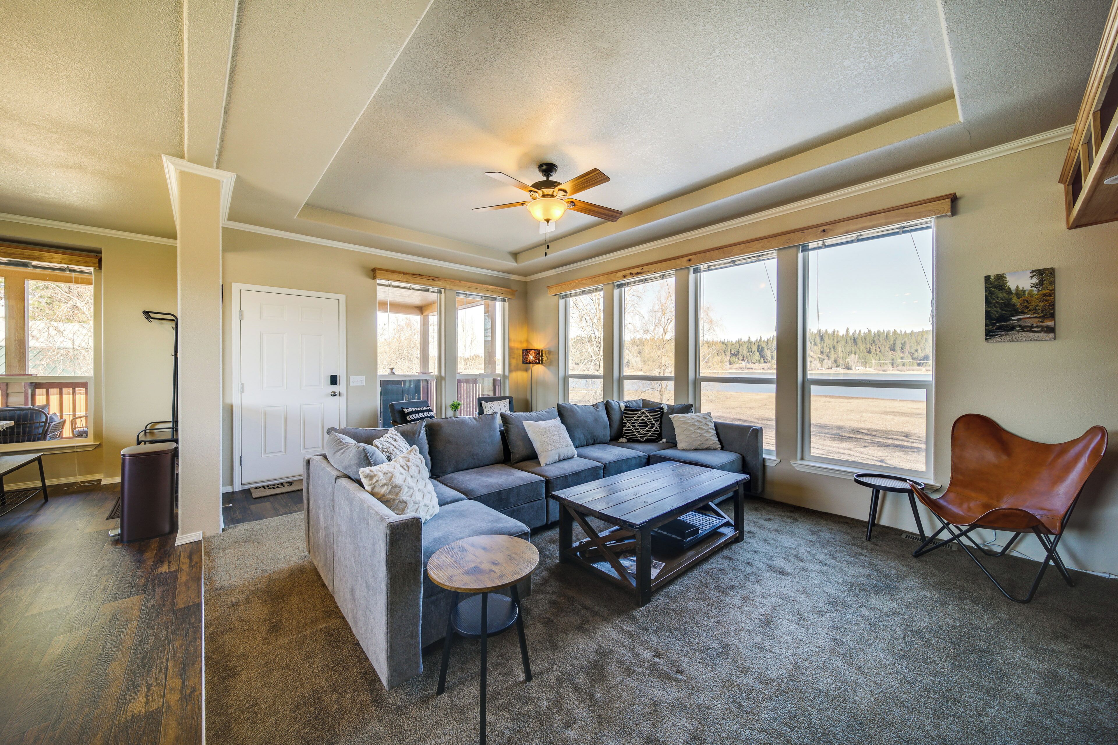 A spacious living room with a sectional sofa, a coffee table, and a ceiling fan. Large windows provide ample natural light and views of the outdoors.