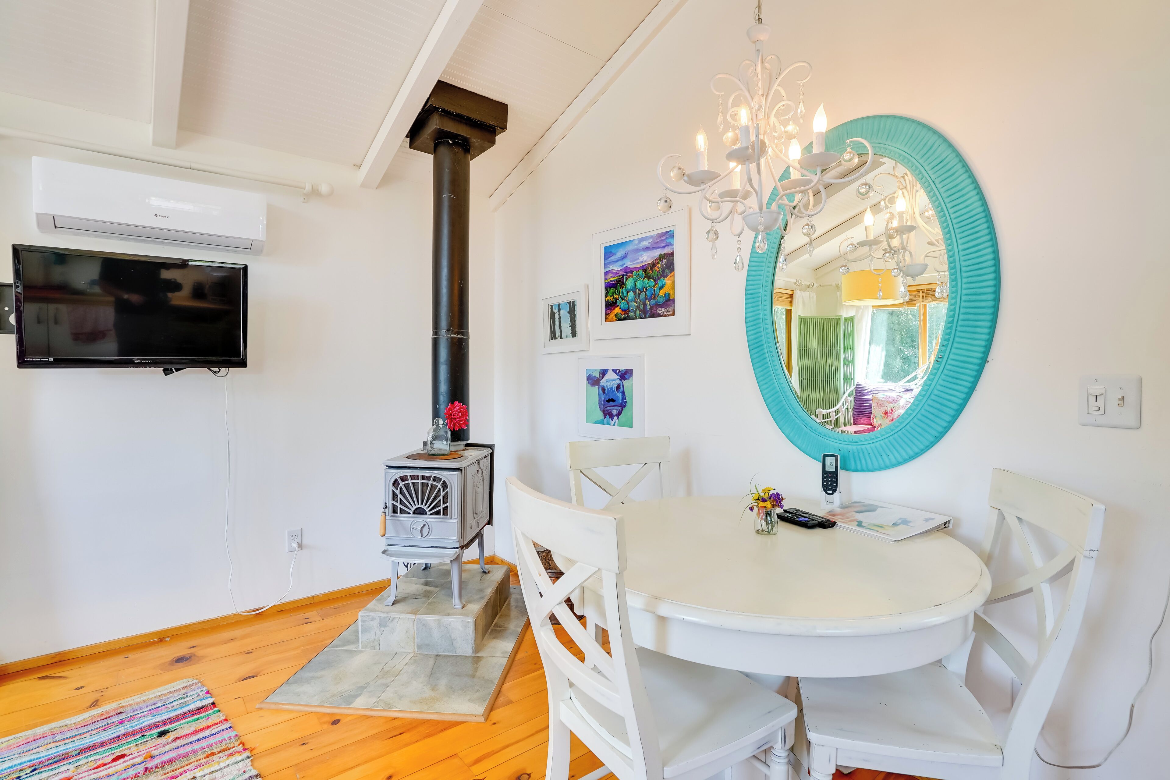 A room with a white wall, a mounted TV, a white table with chairs, a teal-framed mirror, a chandelier, and a wood-burning stove.