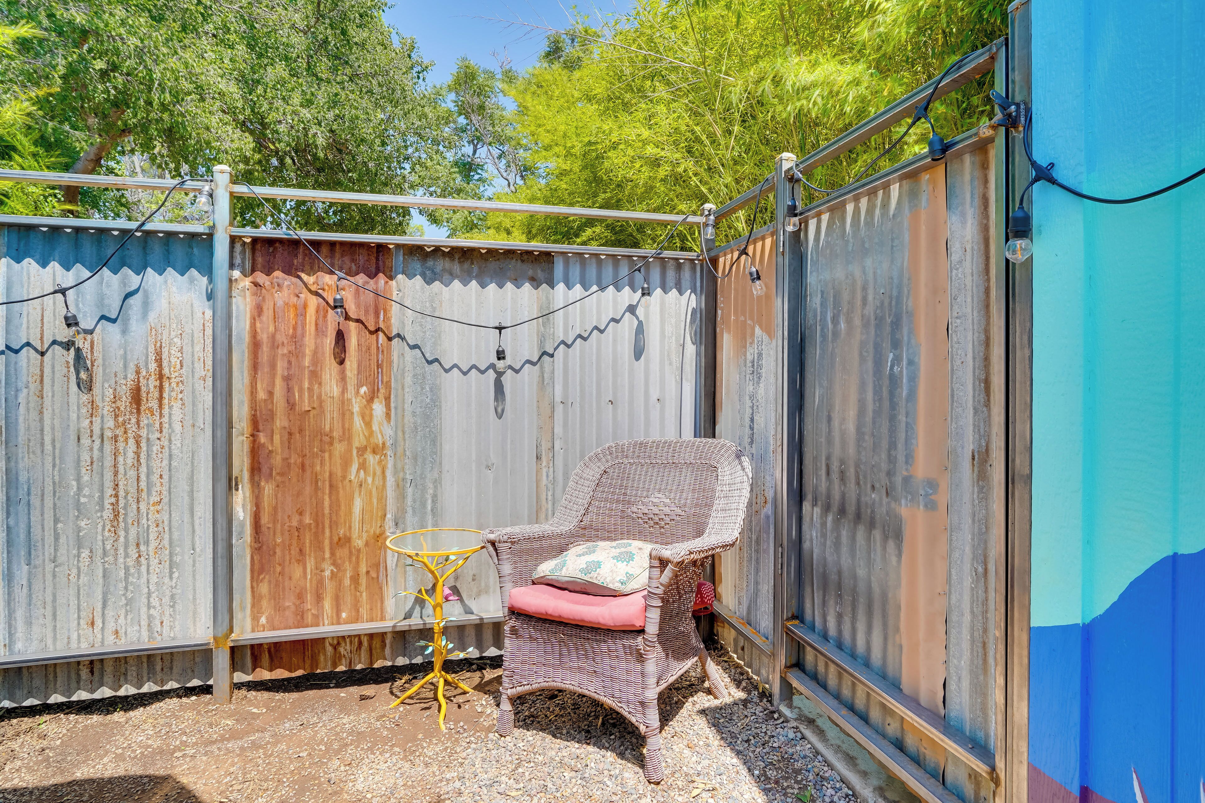 Una silla de mimbre con cojines, una pequeña mesa amarilla y una valla de metal corrugado en un patio trasero.