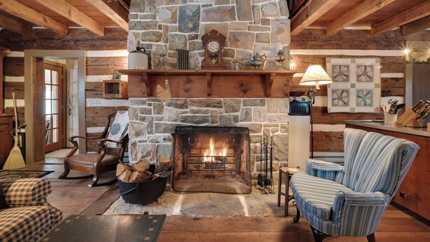 A cozy living room with a stone fireplace, wooden beams, a checkered armchair, a striped armchair, and a dining area with a wooden table and chairs. - Cabin Vacation Rental ~ 8 Mi to Penn State! (Pennsylvania Furnace)
