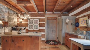 A rustic kitchen with wooden beams, a stone wall, and a large island. - Cabin Vacation Rental ~ 8 Mi to Penn State! (Pennsylvania Furnace)