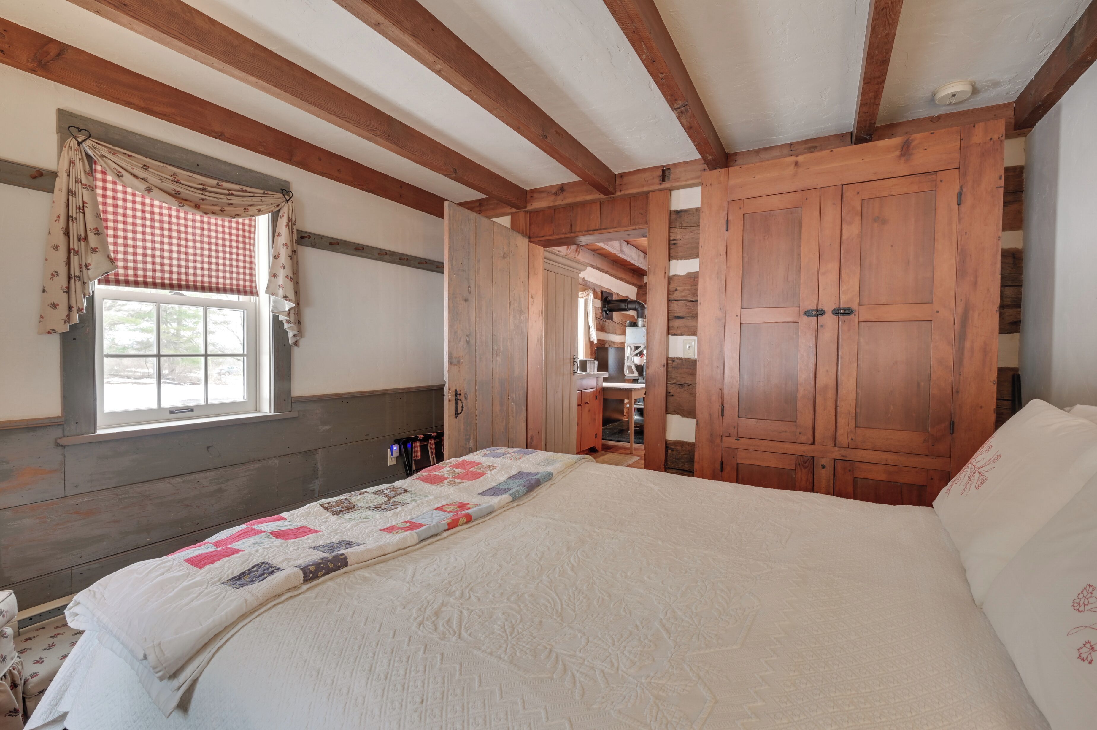 Un dormitorio con una cama grande, vigas de madera, una ventana con cortinas a cuadros y un armario de madera.