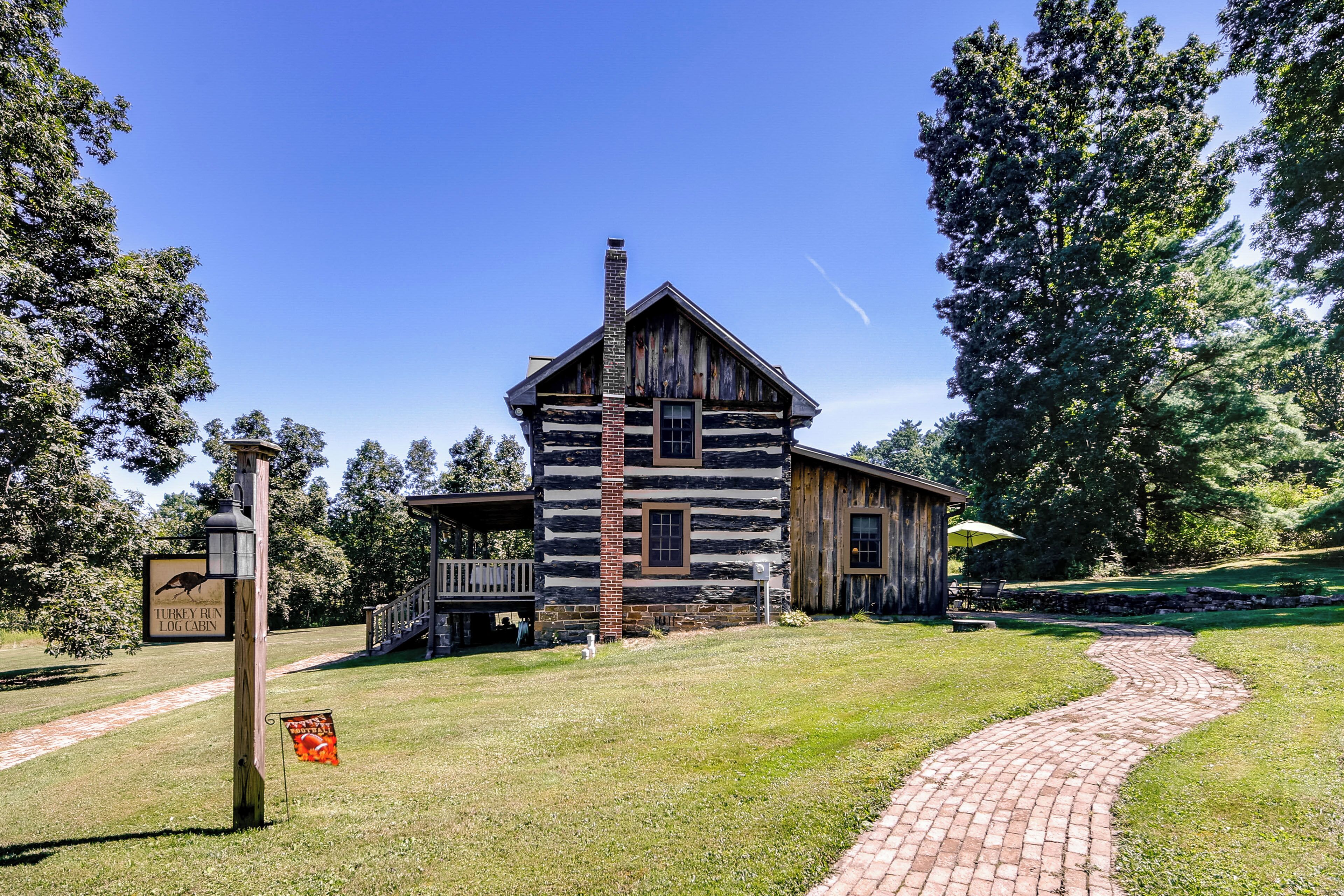 A rustic log cabin with a brick chimney, surrounded by a grassy area and a brick pathway.