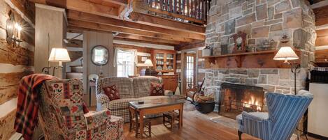 A cozy log cabin interior with a stone fireplace, wooden beams, and a loft area.