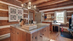 A rustic kitchen with wooden beams, a central island with bar tools, a sink, and a window with curtains. - Cabin Vacation Rental ~ 8 Mi to Penn State! (Pennsylvania Furnace)