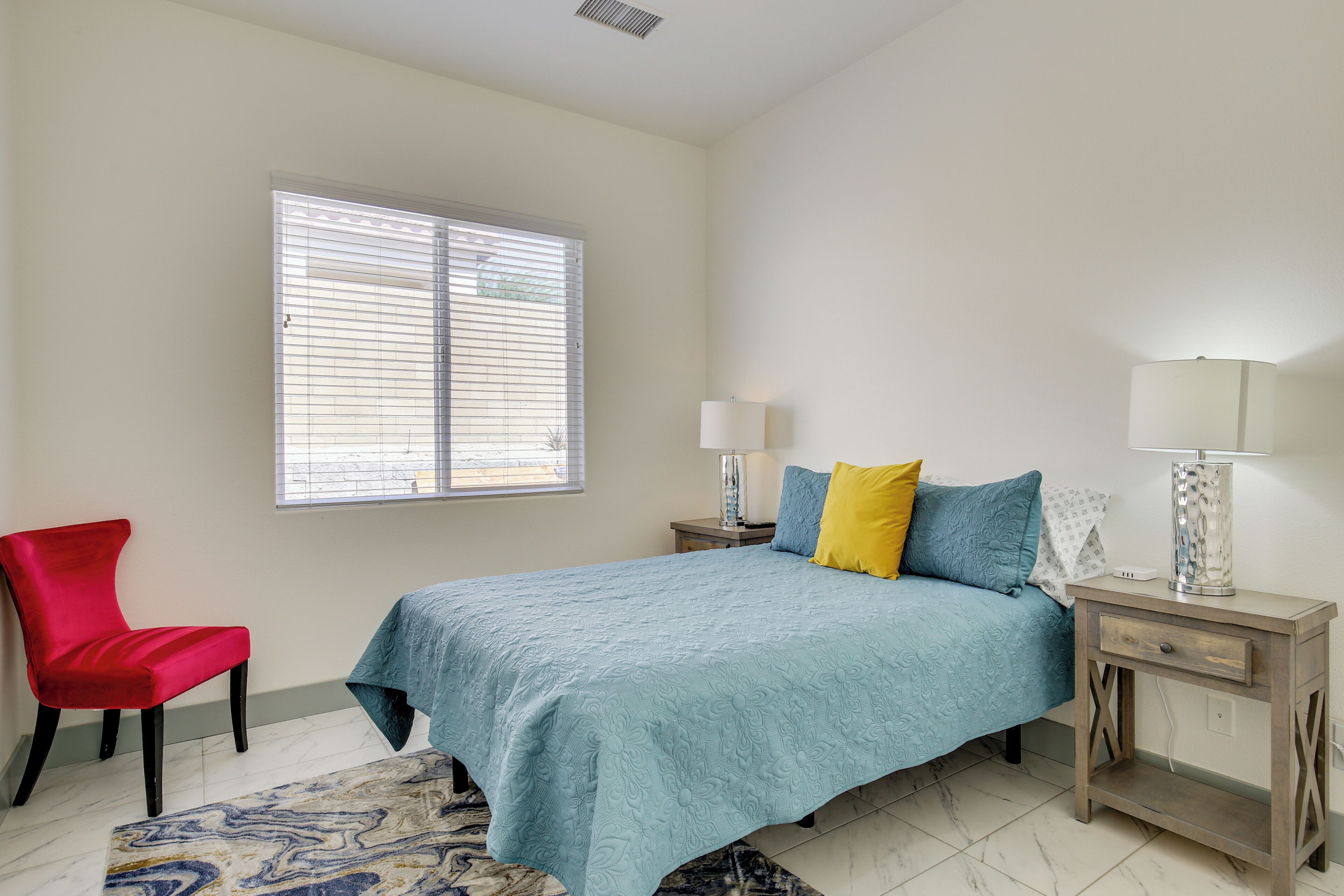 A bedroom with a bed, a nightstand, a lamp, a chair, and a window with blinds.
