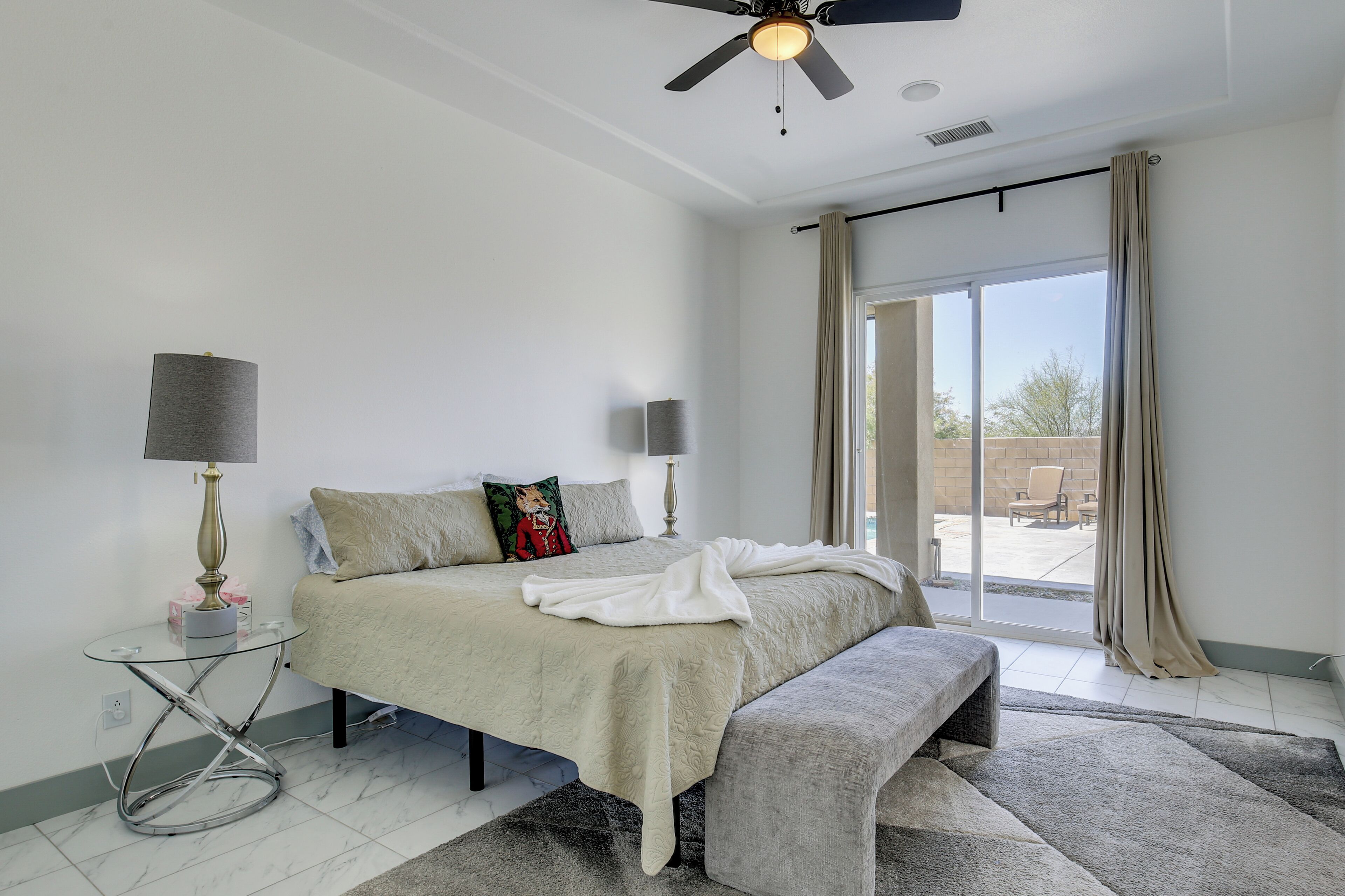 Ein Schlafzimmer mit einem Bett, Nachttischen, einer Bank, einem Deckenventilator und Blick durch eine Schiebetür.