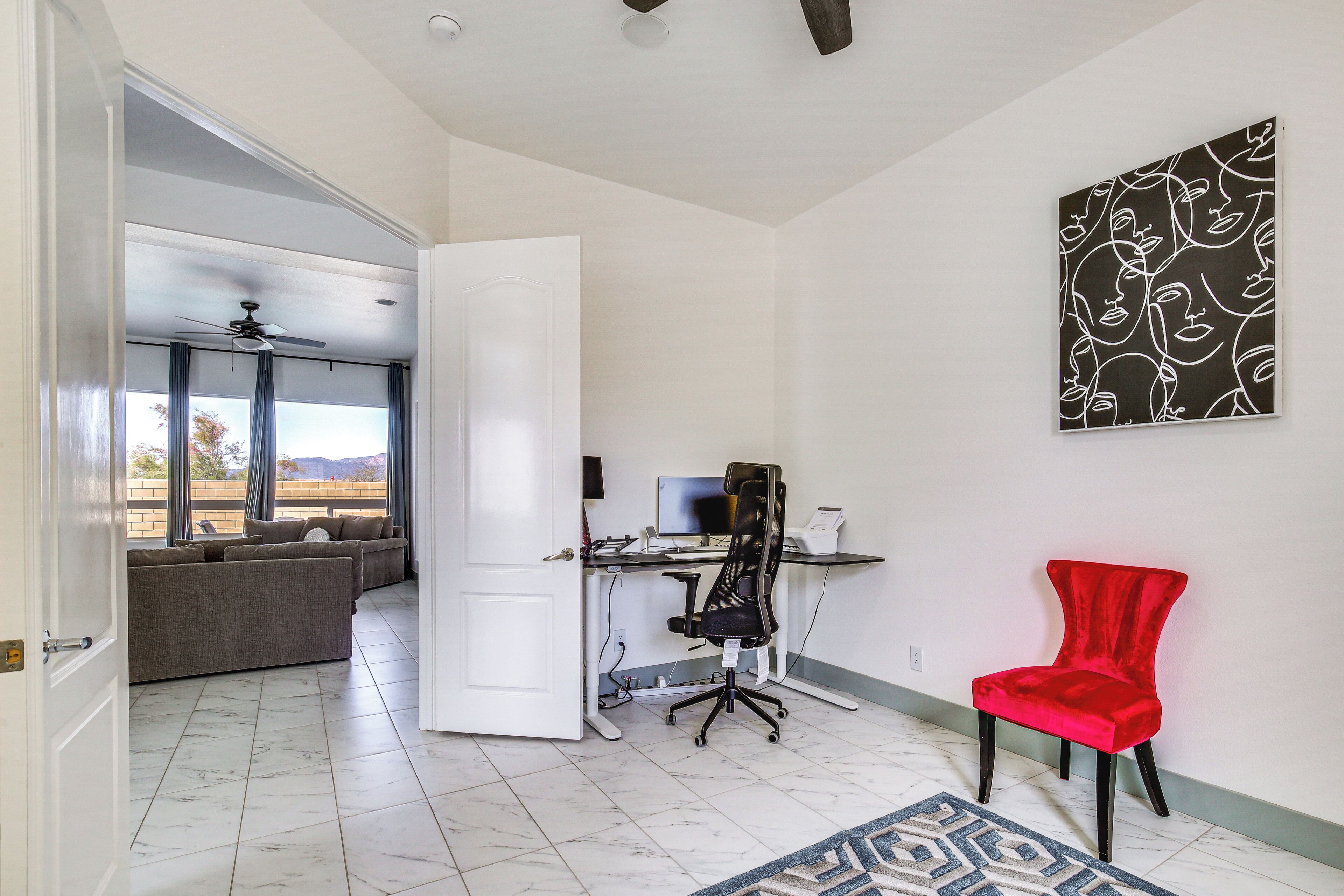 A spacious room with a desk, office chair, and a red chair. There is a large window with curtains, a ceiling fan, and a wall-mounted artwork.
