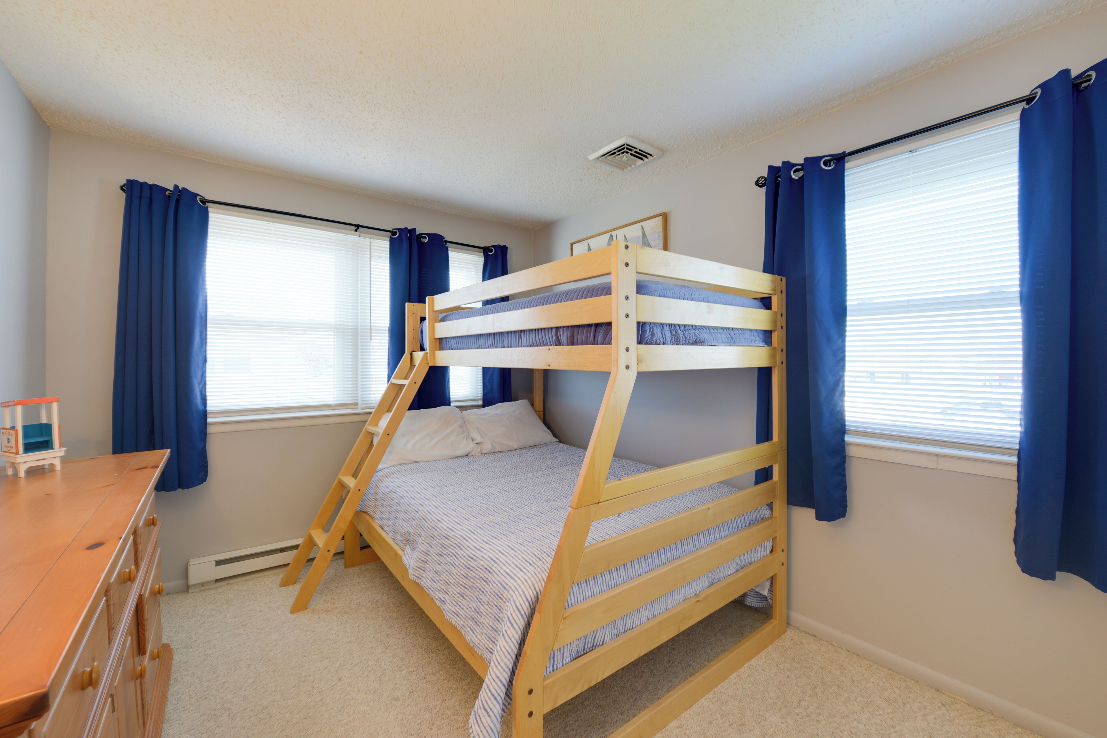A bunk bed in a room with a dresser, windows with blue curtains, and a radiator.