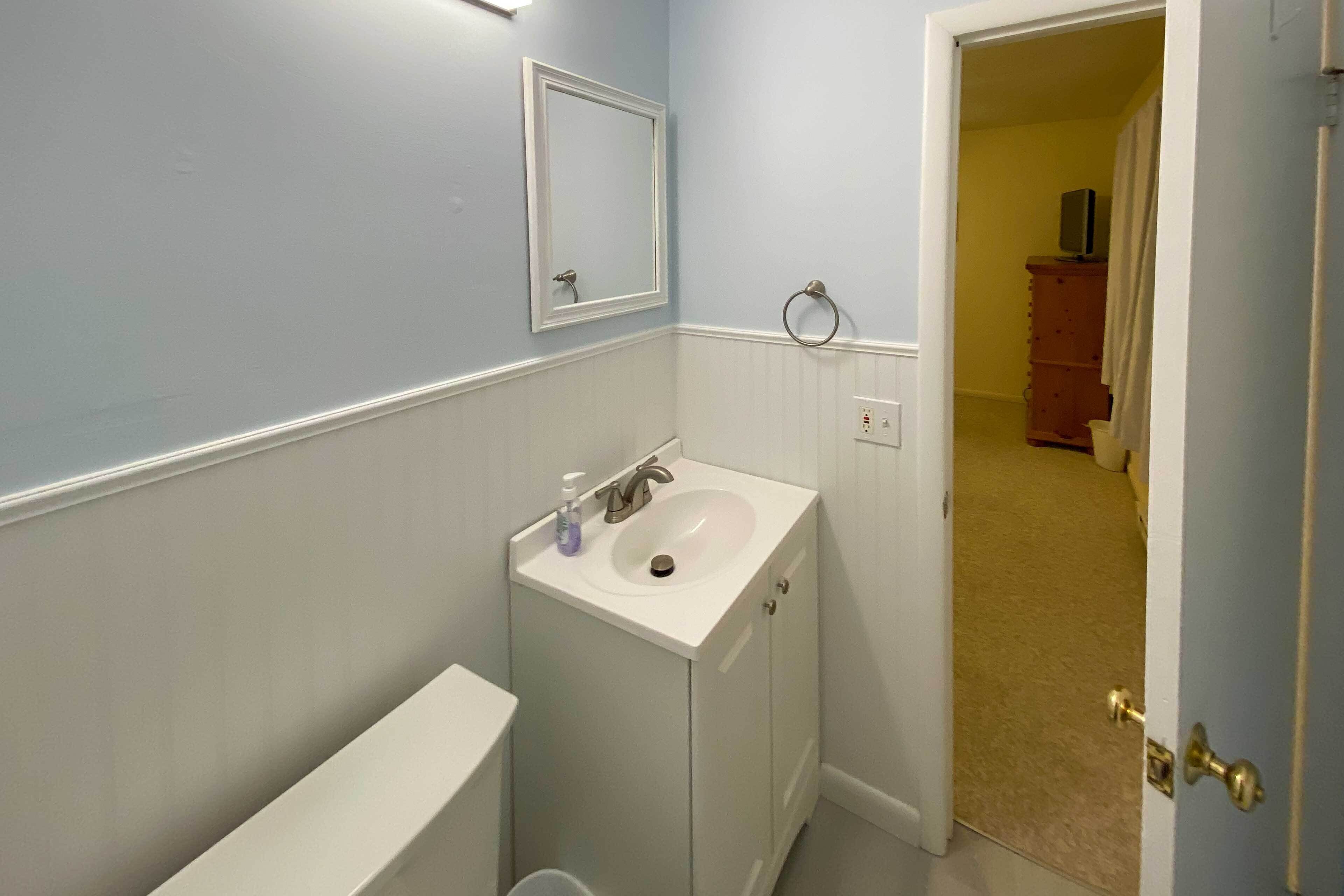Een badkamer met een wit wastafel, een spiegel en een toilet. Er is een deur die naar een andere kamer leidt.