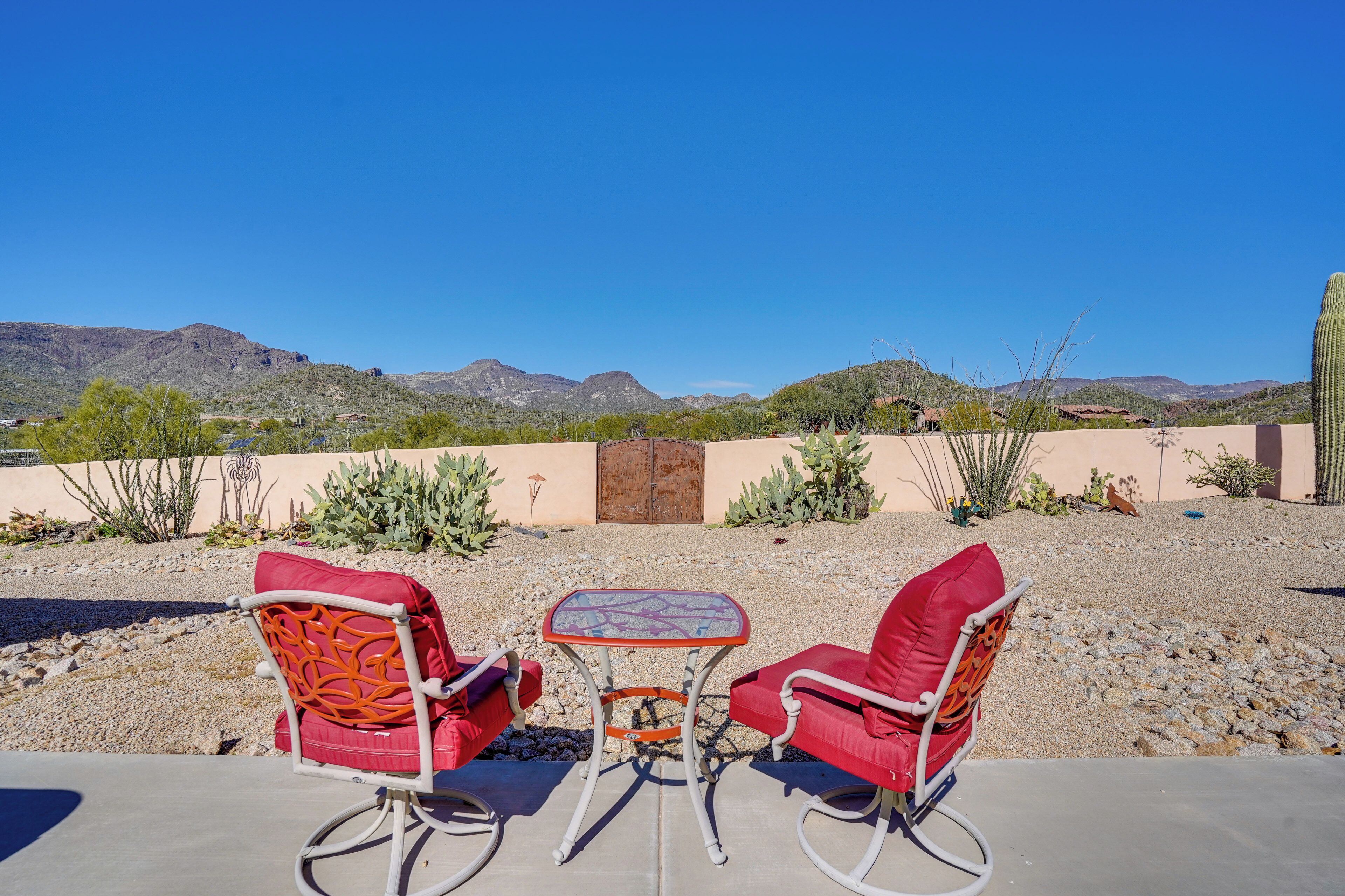 An outdoor seating area with two red chairs and a small table, set against a backdrop of desert landscape and mountains.