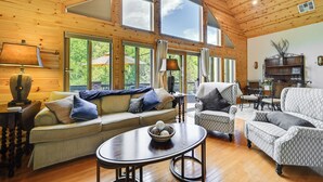 A cozy, well-lit room with a wooden ceiling, large windows, a sofa, armchairs, a coffee table, and a dining area. - Log Cabin in Payson w/ Deck, Mtn & Creek Views (Payson)