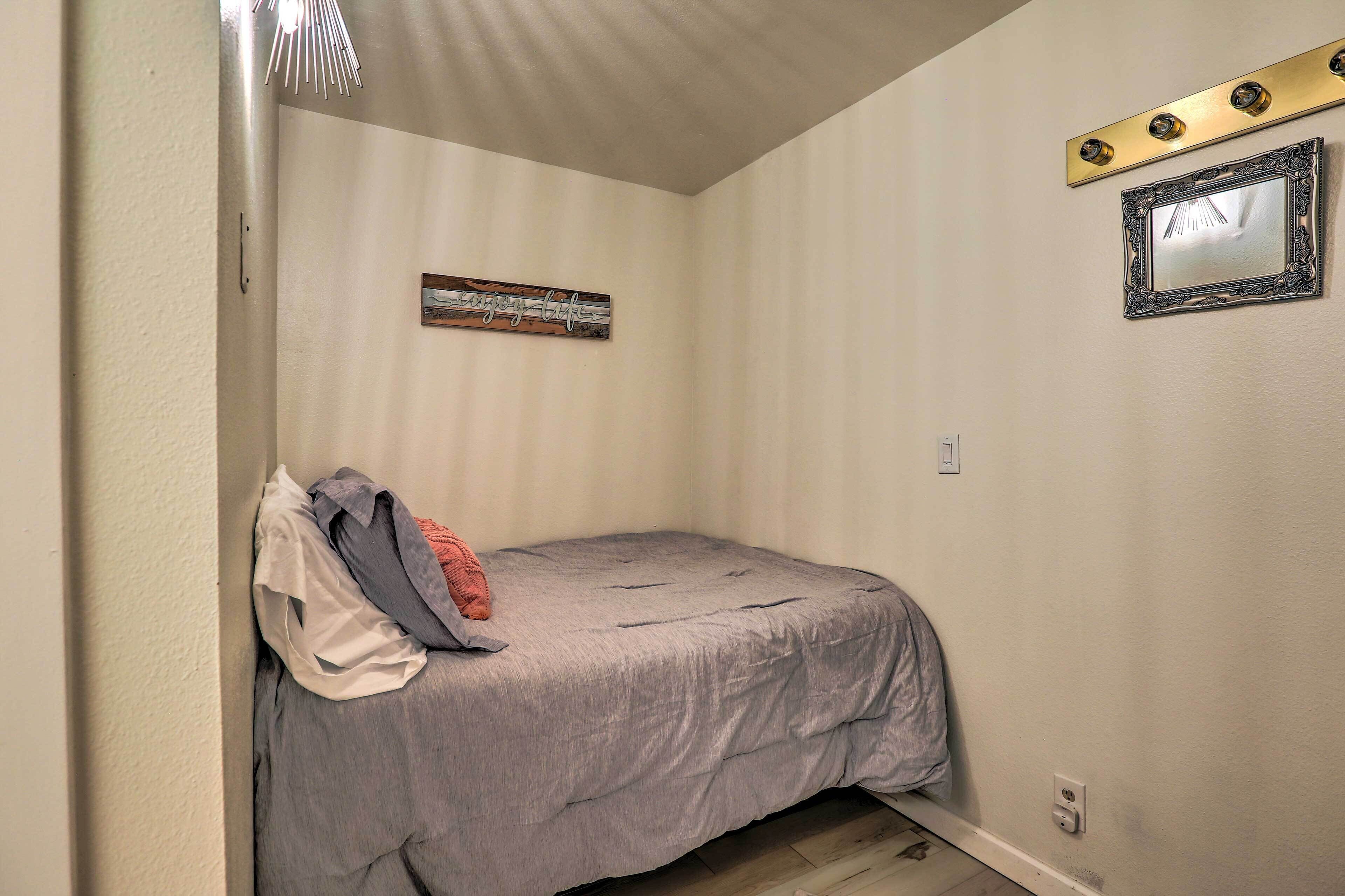 A small, single-bed room with a gray bedspread, a wall-mounted mirror, and a small piece of wall art.
