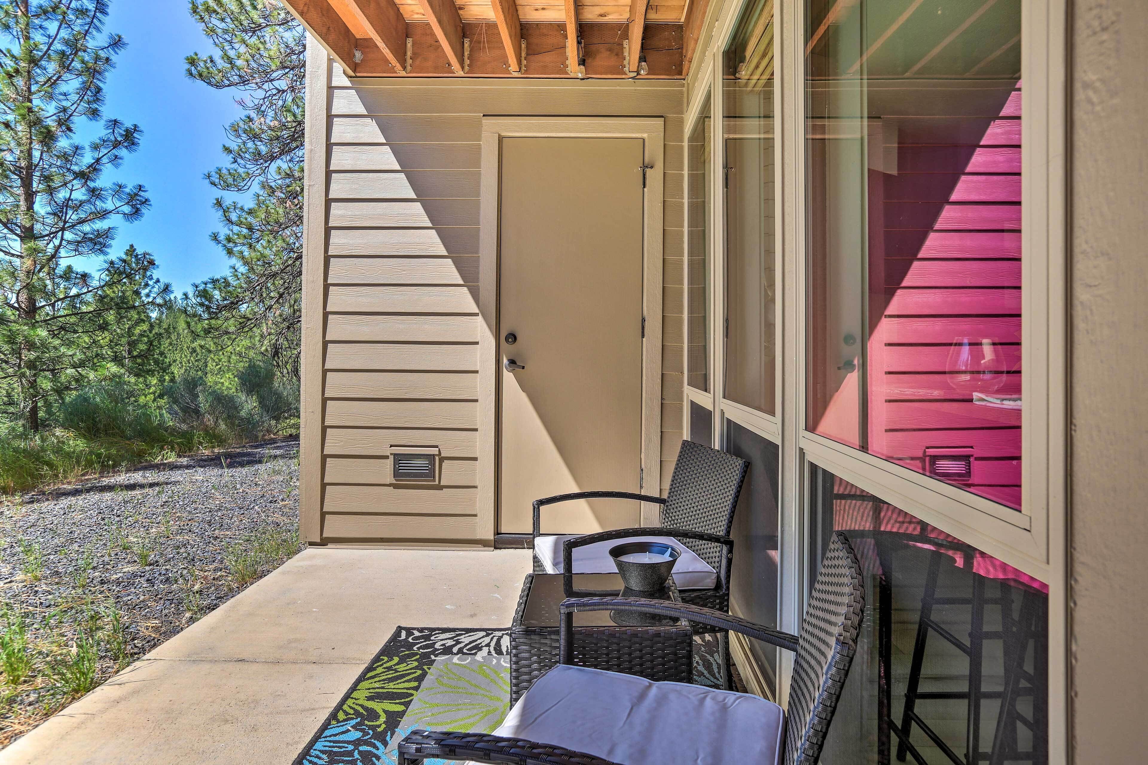 A covered outdoor seating area with a table and chairs, a door, and a view of trees and a building with pink siding.