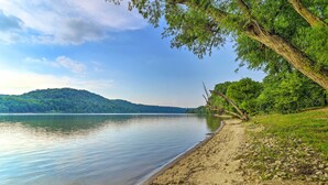 A calm lake with a sandy beach and a forested hillside in the background. - Ohio River Views: Home w/ Fireplace & Deck! (Wolf Creek)