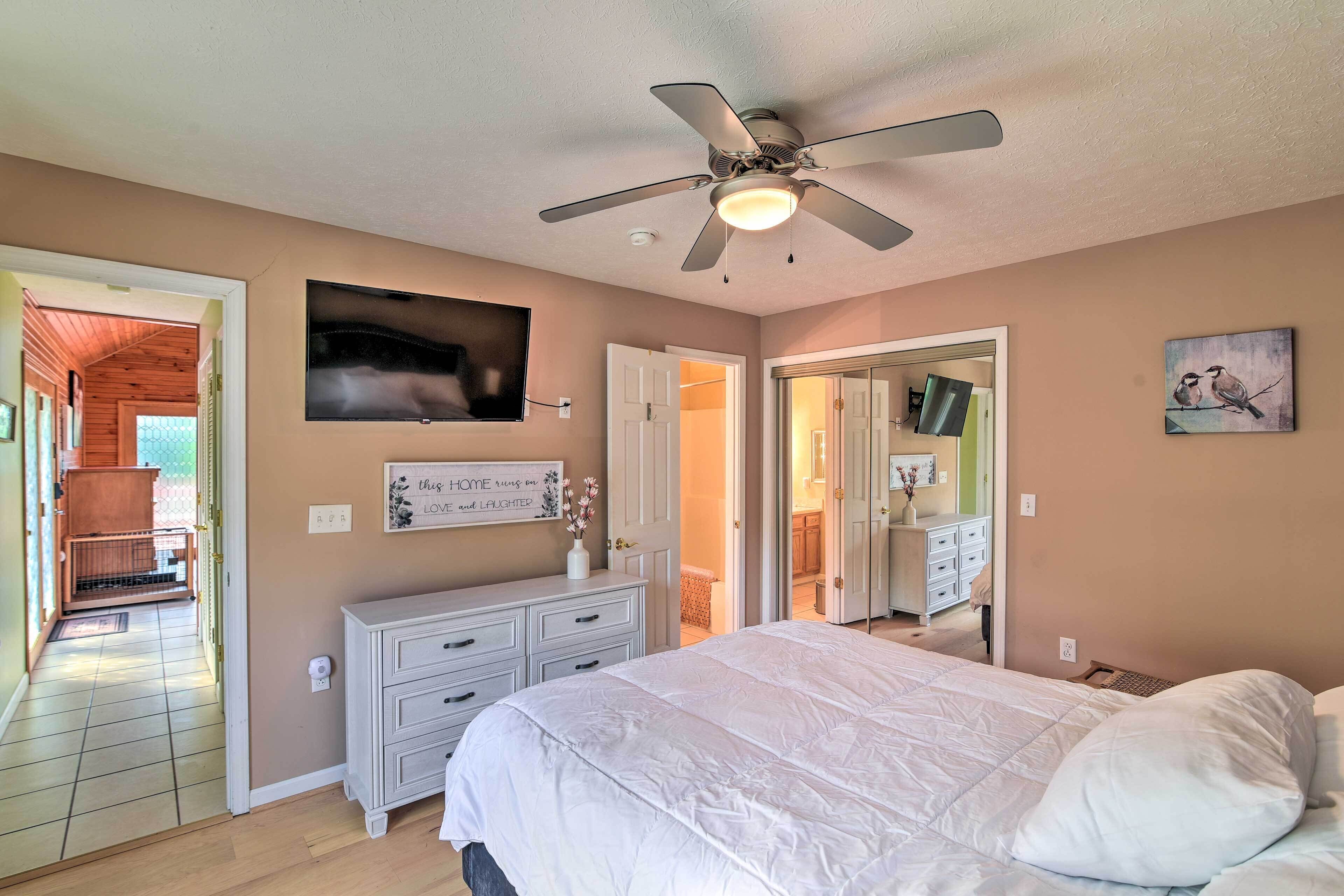 Une chambre à coucher comprenant un lit, une commode, un ventilateur de plafond, un téléviseur et une porte donnant sur une autre pièce.