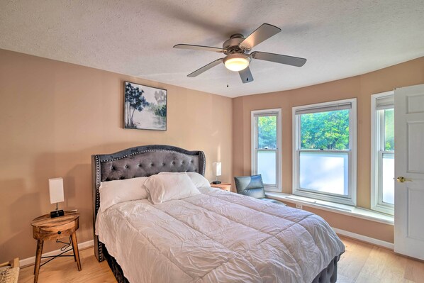 A bedroom with a large bed, a ceiling fan, a window with a view, and a nightstand with a lamp.